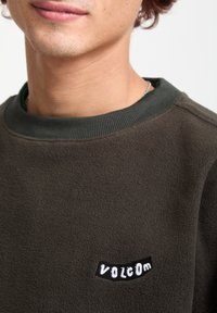 Brązowy polarowy sweatshirt z zielonym ściągaczem w okolicy szyi i małą czarną metką z białą haftowaną logo "Volcom".
