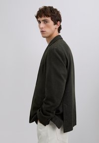 Joven con cabello rizado corto, vestido con una americana verde oscuro y pantalones blancos, de pie de lado con las manos en los bolsillos frente a un fondo liso.