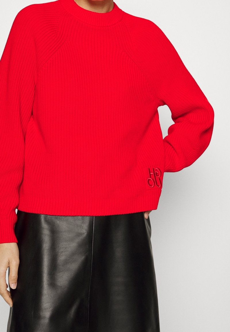 Femme portant un pull côtelé rouge vif avec "HOU" brodé près de l'ourlet, associé à une jupe en cuir noire sur fond blanc.