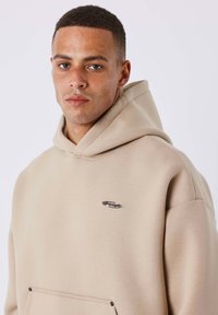 Sudadera con capucha beige hecha de tela suave, con un bolsillo delantero y un pequeño logo decorativo de cadena en el pecho.