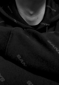 Sudadera negra con una tela texturizada que presenta patrones de logotipo. Vista en primer plano que enfatiza el área del cuello y los detalles de la tela en capas.
