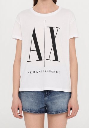 Persoon draagt een wit Armani Exchange T-shirt met grote zwarte "AX" letters en denim shorts, getoond van schouders tot halverwege de dij.