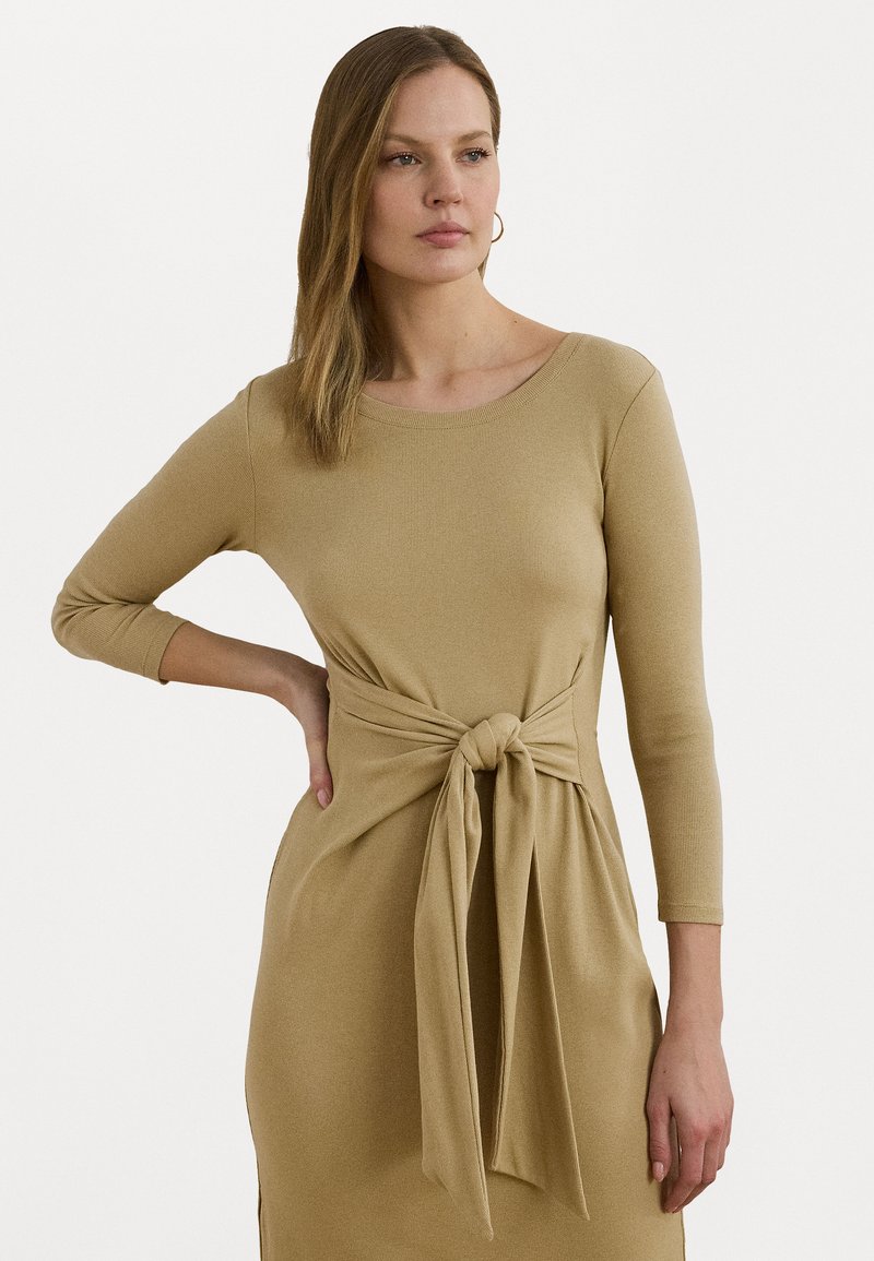 Robe tricotée en beige sable avec un col rond, des manches longues et un accent de lien à l'avant au niveau de la taille. Présente une texture côtelée.