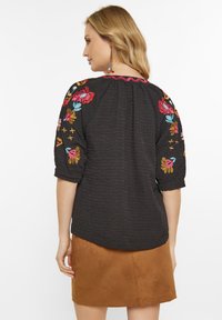 Sort tekstureret bluse med farverig blomsterbroderi på ærmerne og en takkede udskæring. Stylet med en brun ruskindsnederdel.