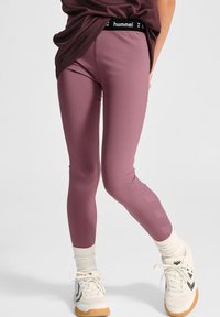 Różowe legginsy z czarnym pasem Hummel, o gładkiej fakturze i geometrycznym wzorze po bokach. Noszone z białymi sneakersami.