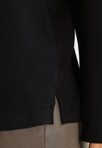 Camisa negra de manga larga con una textura suave y aberturas laterales, que presenta un material en contraste en el dobladillo, combinada con pantalones topo.