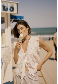 Bijela pamučna bluzica s čipkastim rubom i detaljima od čipke, u kombinaciji s prugastim hlačama. Model drži kornet sladoleda na plaži.
