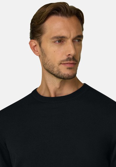 Homme aux cheveux châtain clair et à la barbe, portant un pull noir à col rond, regardant sur le côté sur un fond clair uni.