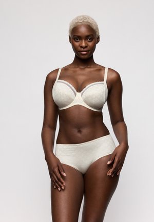 Vrouw met kort blond haar, gekleed in een bijpassende crème kleurige beha en taillehoog slipje met subtiel patroon, staand tegen een effen achtergrond.