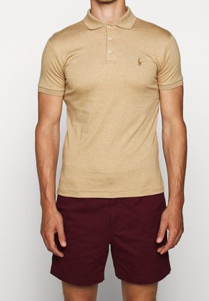 Beige Poloshirt mit Kragen und drei Knöpfen, kurzen Ärmeln und einem kleinen gestickten Logo auf der Brust; kombiniert mit bordeauxroten Shorts.