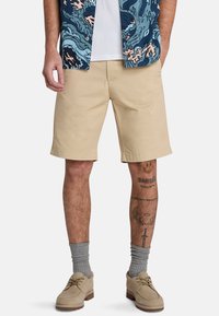 Pantaloni corti di cotone beige con un taglio dritto, tasche laterali e una consistenza liscia. Indossati con calzini grigi e scarpe in suede beige. Camicia a fantasia sopra.