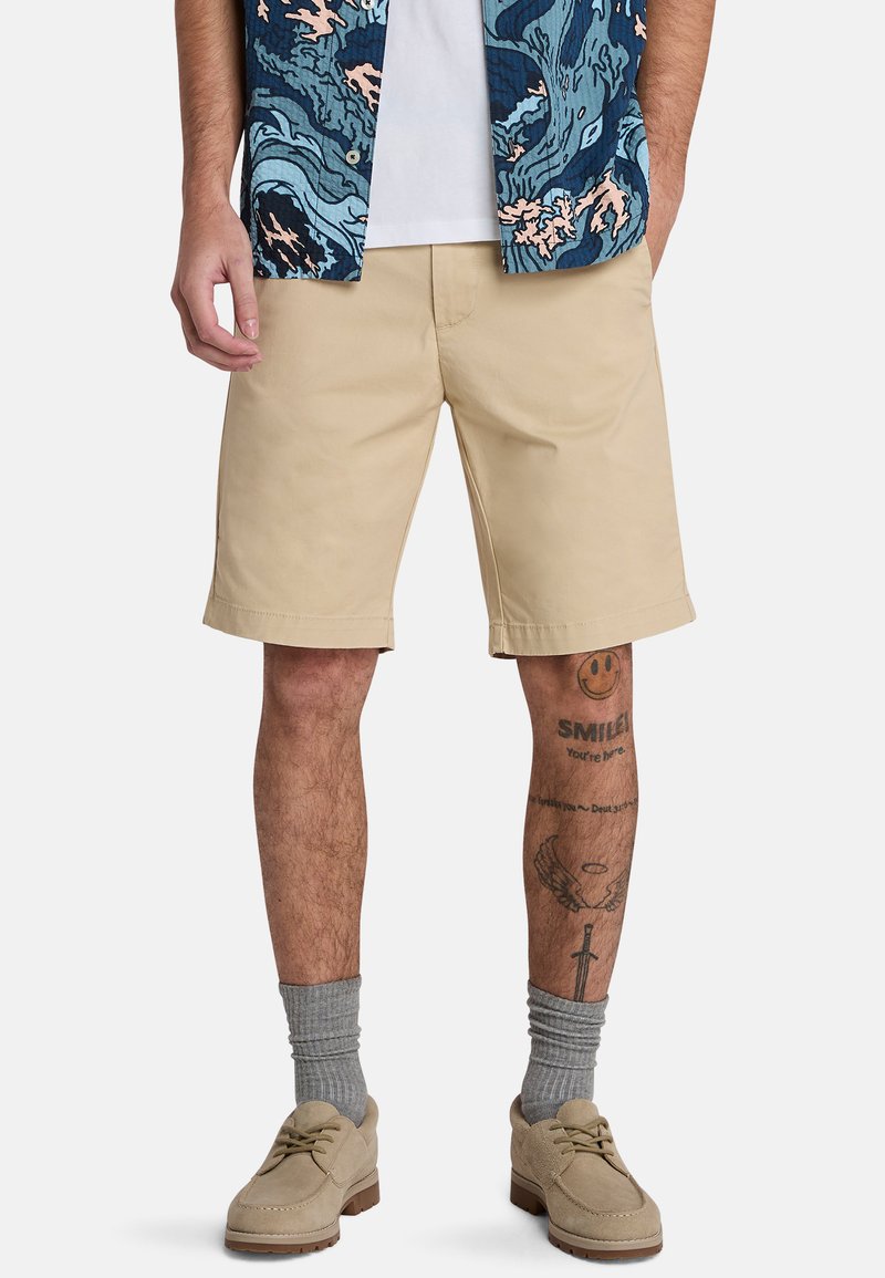 Pantaloni corti di cotone beige con un taglio dritto, tasche laterali e una consistenza liscia. Indossati con calzini grigi e scarpe in suede beige. Camicia a fantasia sopra.