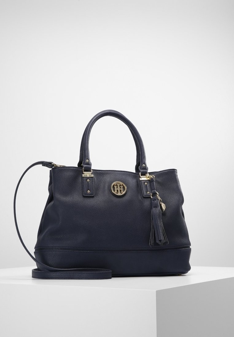 Sac à main en cuir bleu marine avec doubles poignées, bandoulière amovible, emblème de logo doré et breloque pompon sur une surface blanche.