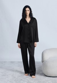 Γυναίκα με μακριά σγουρά μαλλιά, όρθια ξυπόλητη πάνω σε γκρι χαλί, φορώντας φαρδιές μαύρες ριγέ πυτζάμες δίπλα σε μία λευκή υφασμάτινη πολυθρόνα.