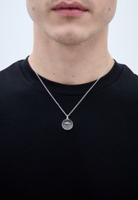 Sudraba kulons, kas veidots apļa formā ar strīpotu saules dizainu, karājas uz delikātas ķēdītes un tiek nēsāts virs melnas krekla.