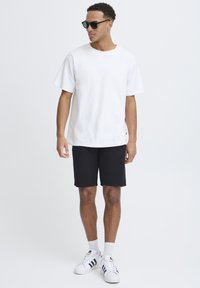 T-shirt en coton blanc, shorts noirs et baskets blanches avec des rayures noires. Modèle portant des lunettes de soleil foncées, debout contre un fond neutre.