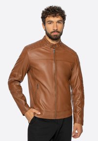 Muž s vousy v hnědé kožené bundě na zip s prošívanými detaily na ramenech, stojící s jednou rukou v kapse před jednobarevným pozadím.