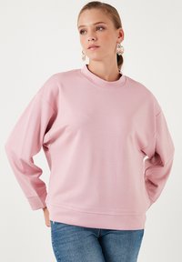 Maglione a maniche lunghe rosa chiaro con colletto alto, texture morbida, spalle cadenti e polsini a coste. Indossato con jeans in denim blu.