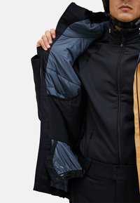 Giacca nera isolante con una fodera liscia blu, dotata di chiusura con zip, tasche laterali e un pannello marrone a contrasto. Tessuto dalla trama visibile.