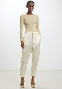 Top a maniche lunghe beige a costine con un design aderente, abbinato a pantaloni cargo in satin crema con tasche laterali e polsini elastici. Tacchi alti dorati.