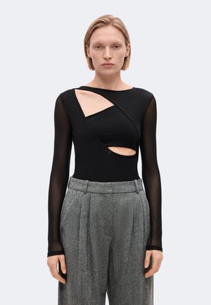 Femme aux cheveux blonds courts portant un haut noir avec des découpes et des manches longues transparentes, associé à un pantalon gris taille haute à jambes larges.
