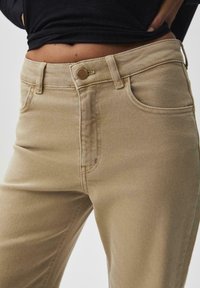 Beige Baumwolljeans mit hoher Taille, ausgestattet mit Fronttaschen, einem Metallknopfverschluss und Ton-in-Ton-Nähten. Glattes, leicht strukturiertes Material.