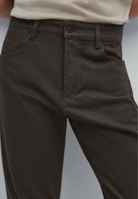 Pantaloni scuri in denim di colore marrone scuro, dotati di cinque tasche, chiusura con bottone e gamba dritta. Il tessuto presenta una leggera trama.
