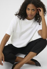 Witte T-shirt met korte mouwen en een ronde hals, gecombineerd met zwarte cropped broeken. Het model heeft krullend haar en draagt zwarte ballerina's.