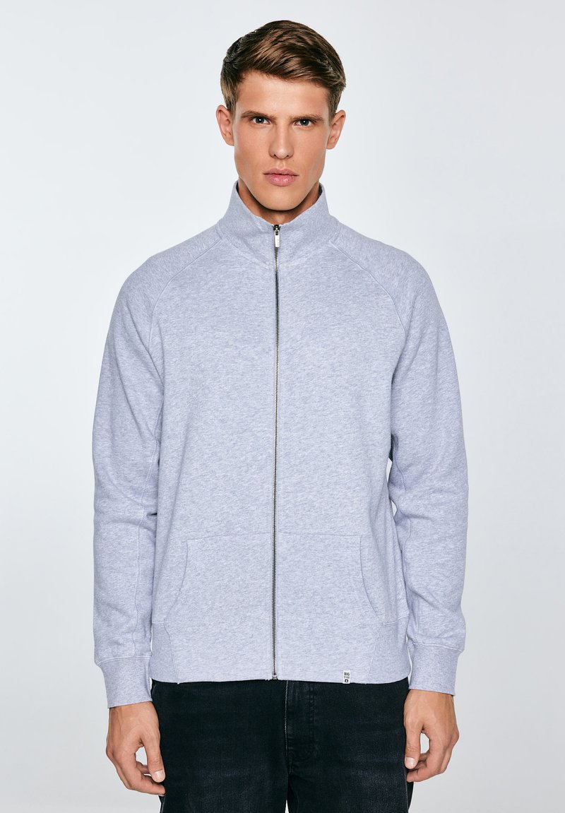 Młody mężczyzna w jasnoszarej bluzie na zamek z wysokim kołnierzem i przednimi kieszeniami, stojący na tle jednolitego jasnego tła.