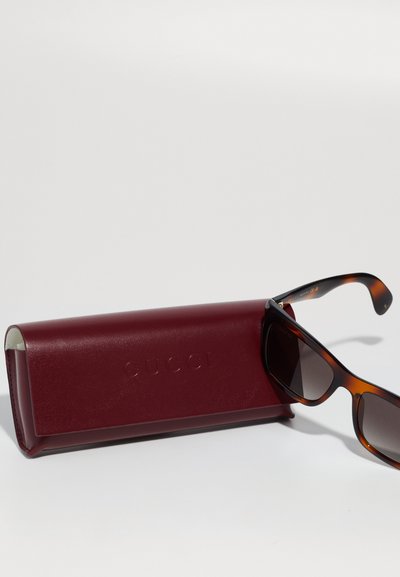 Gucci sončni očala v rjavi kornjačevini z temnimi lečami, postavljeni ob strani maroon usnjene torbice z vtisnjenim logotipom.