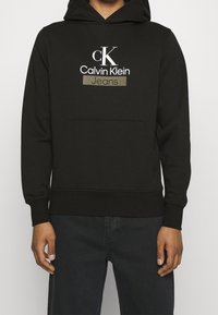 Person som bär en svart Calvin Klein-hoodie med framficka och logotexten "cK Calvin Klein Jeans" på bröstet, kombinerad med mörka byxor.