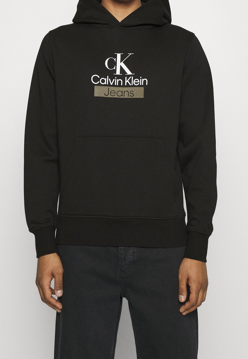 Person som bär en svart Calvin Klein-hoodie med framficka och logotexten "cK Calvin Klein Jeans" på bröstet, kombinerad med mörka byxor.