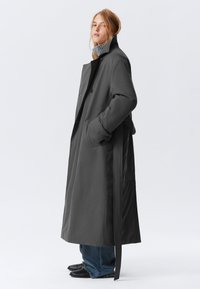Cappotto lungo grigio con colletto alto, due tasche frontali e cintura. Tessuto liscio e design strutturato. Indossato su un maglione grigio e jeans.