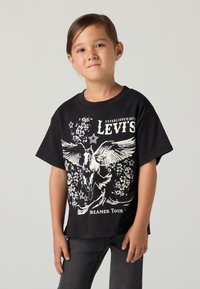 Noor brünettidega laps, kes kannab musta Levi's T-särki, millel on valge Pegasus ja tekst "Dreamer Tour", kombineerituna tumedate teksadega.