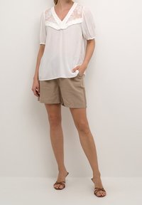 Blusa blanca con acentos de encaje transparente y detalle con volantes en el escote, combinada con pantalones cortos beige y sandalias de tiras marrones.