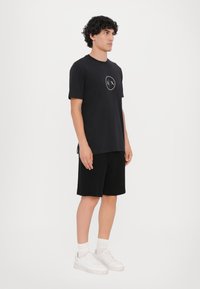 Sort t-shirt med et rundt logo i hvid, parret med sorte shorts. Modellen har hvide sneakers og ankelstrømper på, med krøllet hår.