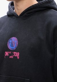 Gros plan sur une personne portant un sweat à capuche noir avec un graphisme violet et rose représentant un visage souriant stylisé et le texte « ON TOUR SUPPORT » sur la poitrine.