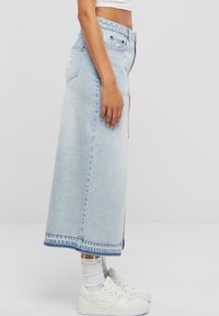 Jupe maxi en denim bleu clair avec fentes sur les côtés, coutures contrastées et une étiquette de marque à l'arrière. Associée à des baskets blanches.