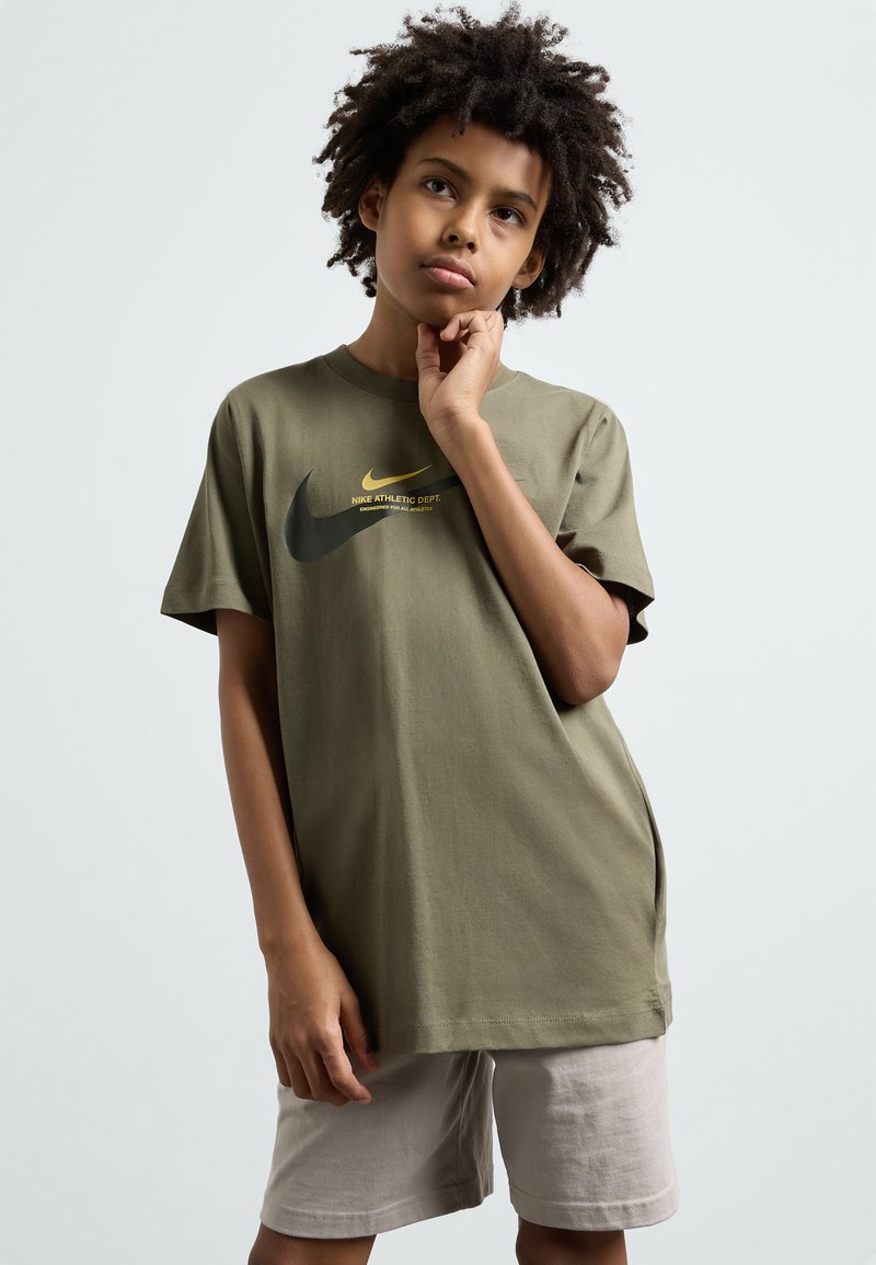Olive žalios spalvos medvilninis marškinėlis su apvali aptempta priekiu, trumpomis rankovėmis, priekyje su juodu ir geltonu Nike logotipo tekstu. Šviesūs šortai.