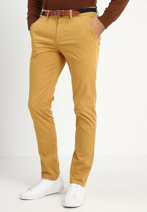 Hombre con pantalones ajustados color mostaza con cinturón de cuero marrón, camisa marrón de manga larga y zapatillas blancas, con las manos en los bolsillos.