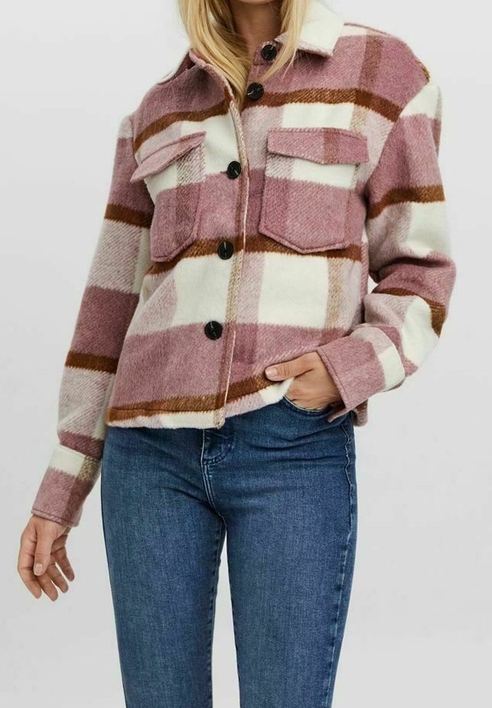 Femme portant une veste-chemise à carreaux rose, blanche et marron boutonnée avec deux poches poitrine et un jean bleu, une main dans la poche.