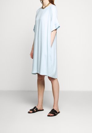 Femme portant une robe ample bleu clair mi-longue à manches courtes et des sandales noires à enfiler, debout devant un fond clair uni.