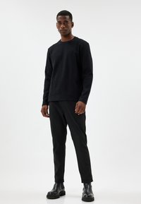 Langärmeliges schwarzes geripptes Strickoberteil, kombiniert mit schwarzen, schmal zulaufenden Hosen. Das Modell trägt schwarze Stiefeletten und steht vor einem neutralen Hintergrund.