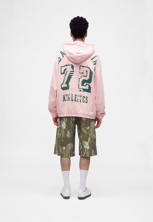 Mann trägt einen pinken Hoodie mit dem Text "72 Athletics" auf dem Rücken, grüne Shorts mit Blattmuster, weiße Socken und weiße Sneakers, steht mit dem Rücken zugewandt.
