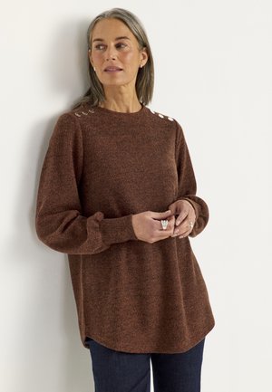 Kvinde med gråt hår iført en brun strikket sweater med skulderknapper og mørke bukser, stående ved en hvid væg og kigger til siden.