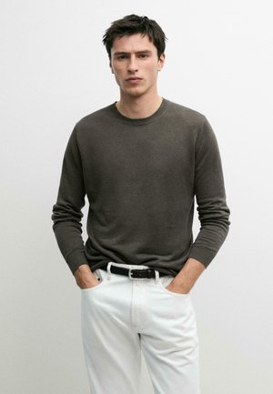 Młody mężczyzna o ciemnych włosach nosi oliwkowy sweter z długim rękawem włożony do białych spodni z czarnym paskiem, ręce w kieszeniach, neutralne tło.