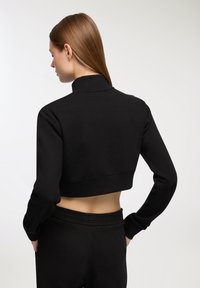 Felpa nera corta con colletto alto e maniche lunghe. Tessuto morbido con un design aderente e polsini a costine, indossata con pantaloni coordinati.