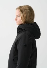 Zwarte waterdichte jas met een capuchon, met een gladde textuur en een logopatch op de mouw. Er zijn ritssluitingen aan de zijkant.