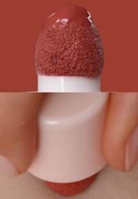 Aplicador cosmético con punta de esponja con un líquido marrón rojizo oscuro en la parte superior, sostenido contra la piel, con una base cilíndrica blanca.