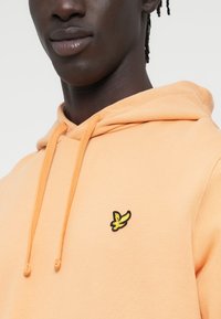 Πορτοκαλί φούτερ με κουκούλα και κορδόνια, με κεντημένο λογότυπο σε μαύρο και κίτρινο στην αριστερή πλευρά του στήθους. Μαλακό, υφή βαμβακερό υλικό.